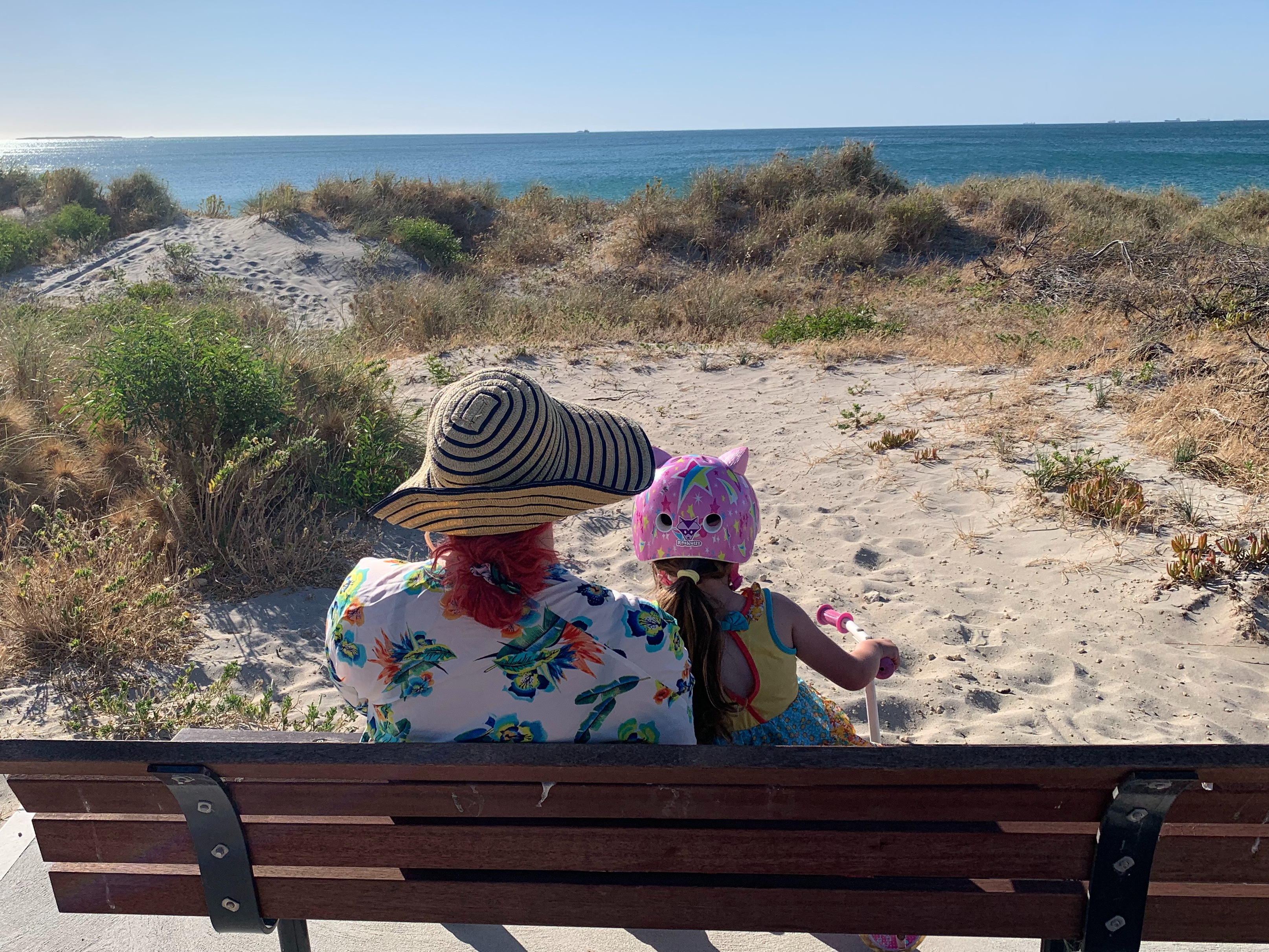 woman with stripy hat, floral shirt and red hair facing away on a bench, a young girl with long brown hair and a pink helmet next to her, both looking at the ocean which is visible over sand and grasses